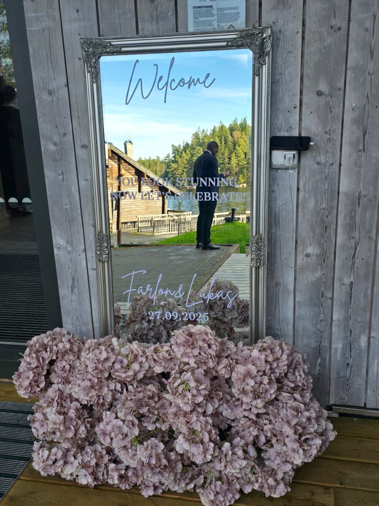 A decorative mirror framed in ornate silver, displaying 'Welcome' and 'You look stunning. Now let's celebrate!' text. A man in formal attire is reflected in the mirror, standing beside a floral arrangement of pink blossoms at the base.