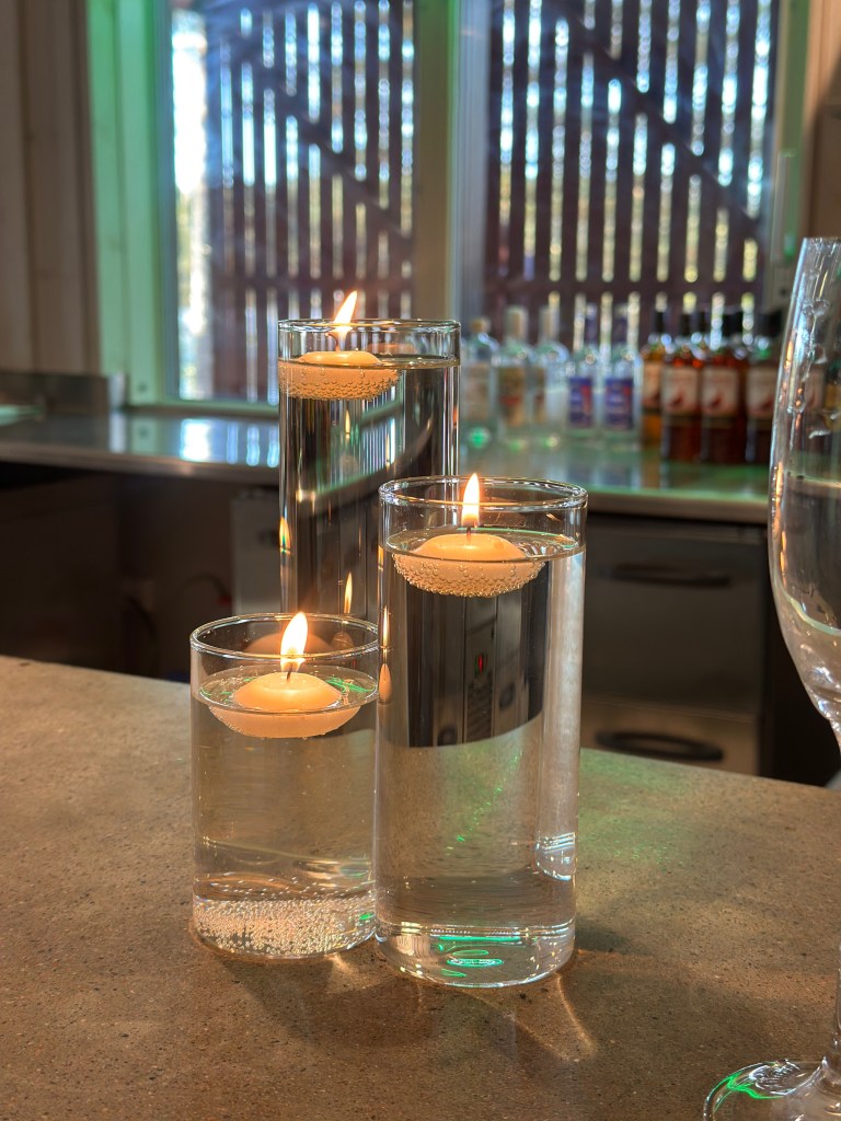 Three glass candle holders filled with water, each containing a floating candle, on a countertop with blurred background.