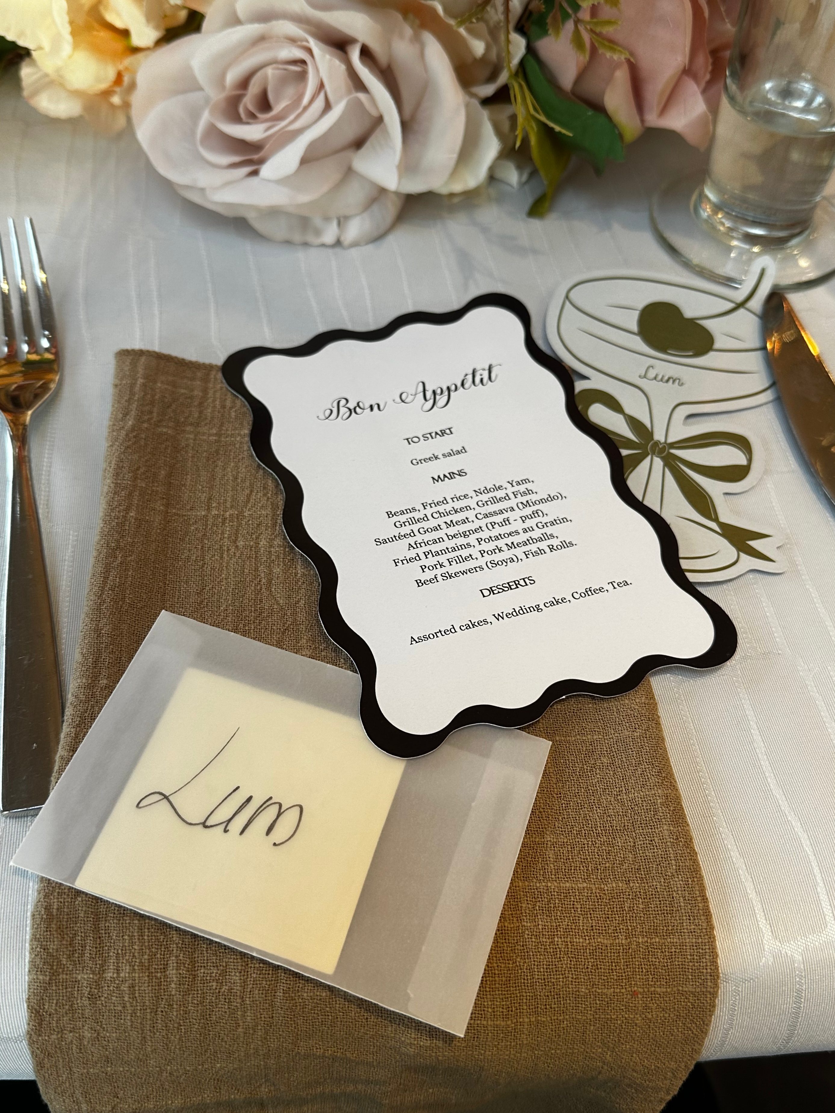 Table setting featuring a menu card titled 'Bon Appétit' with a floral arrangement in the background, and a name card labeled 'Lum' on a beige napkin.