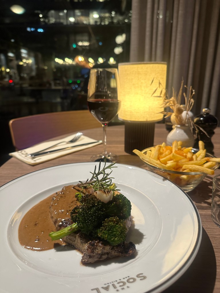 A plate of grilled steak topped with broccoli and garnished with rosemary, served with a rich sauce, accompanied by a side of French fries and a glass of red wine, with a warm lamp in a softly lit restaurant setting.