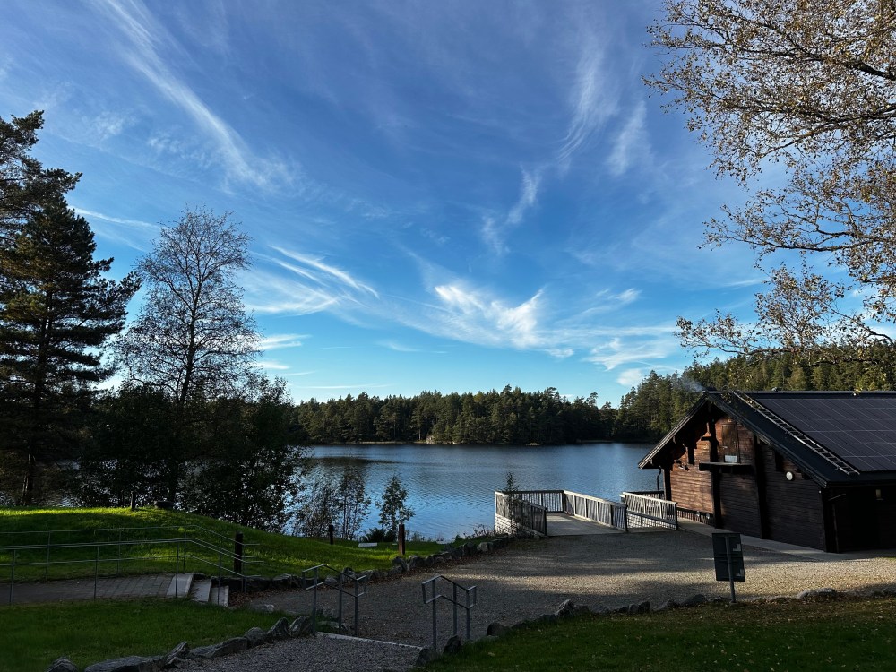 Scenic view of a tranquil lake surrounded by trees under a blue sky with clouds, featuring a wooden building with solar panels and a pathway leading down to the water.
