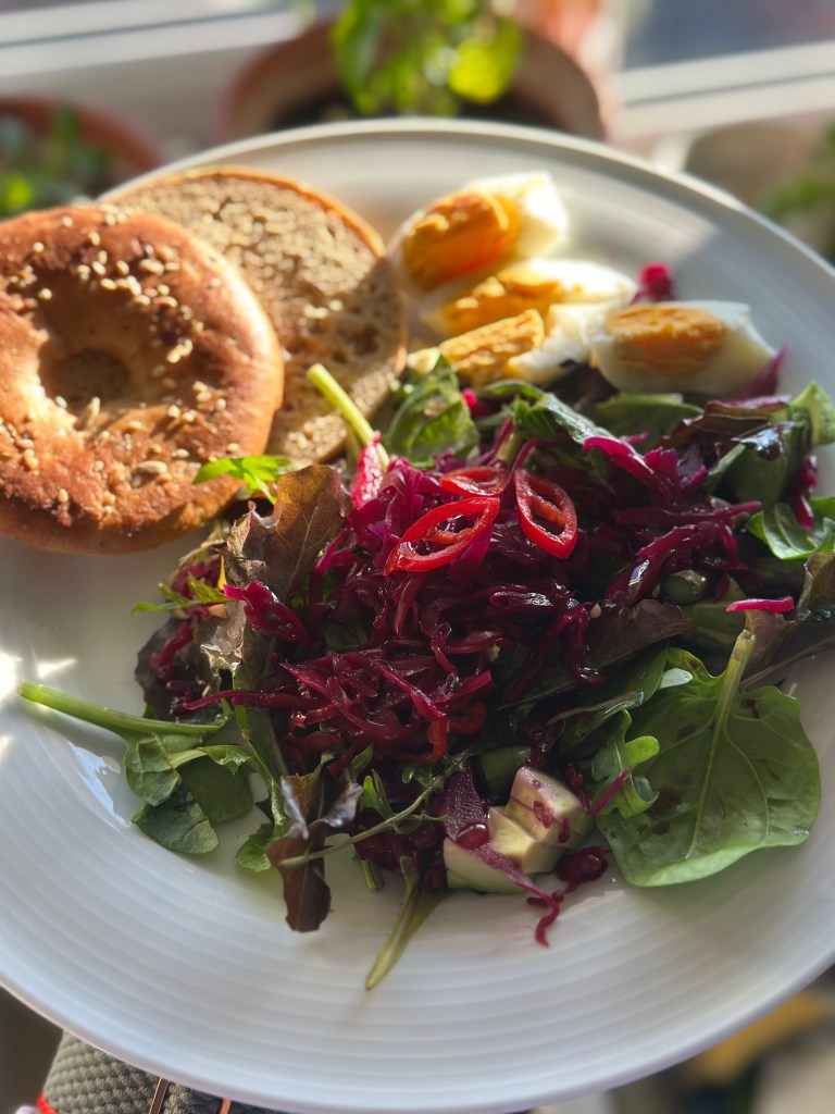 A plate featuring a colorful salad with mixed greens, red sauerkraut, and diced avocados, alongside a sesame seed bagel and boiled eggs.
